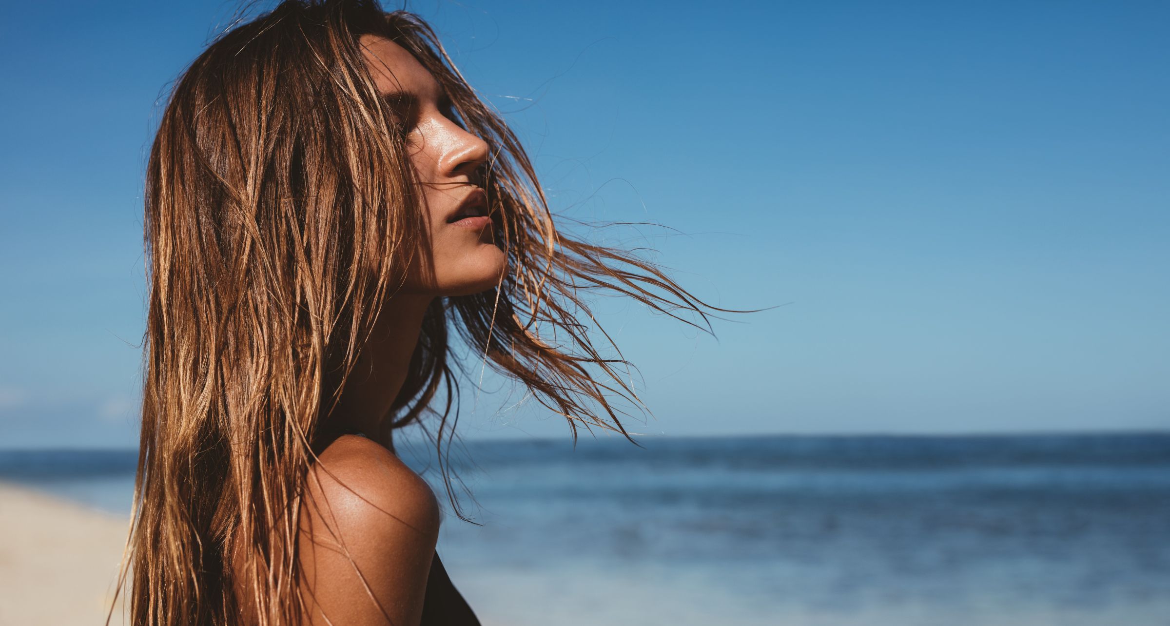 Donna in spiaggia di profilo con capelli al vento e il mare di sfondo.